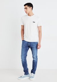 Camiseta blanca de algodón con el logotipo negro de "Lee", combinada con jeans de mezclilla azul y zapatos de color azul claro. Ajuste slim, de manga corta, estilo casual.