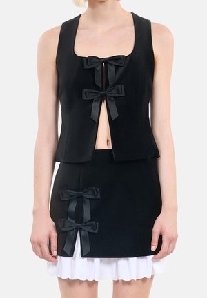 Mujer con un conjunto de top negro sin mangas y falda, ambos con lazos negros decorativos, con un dobladillo blanco con volantes visible bajo la falda.