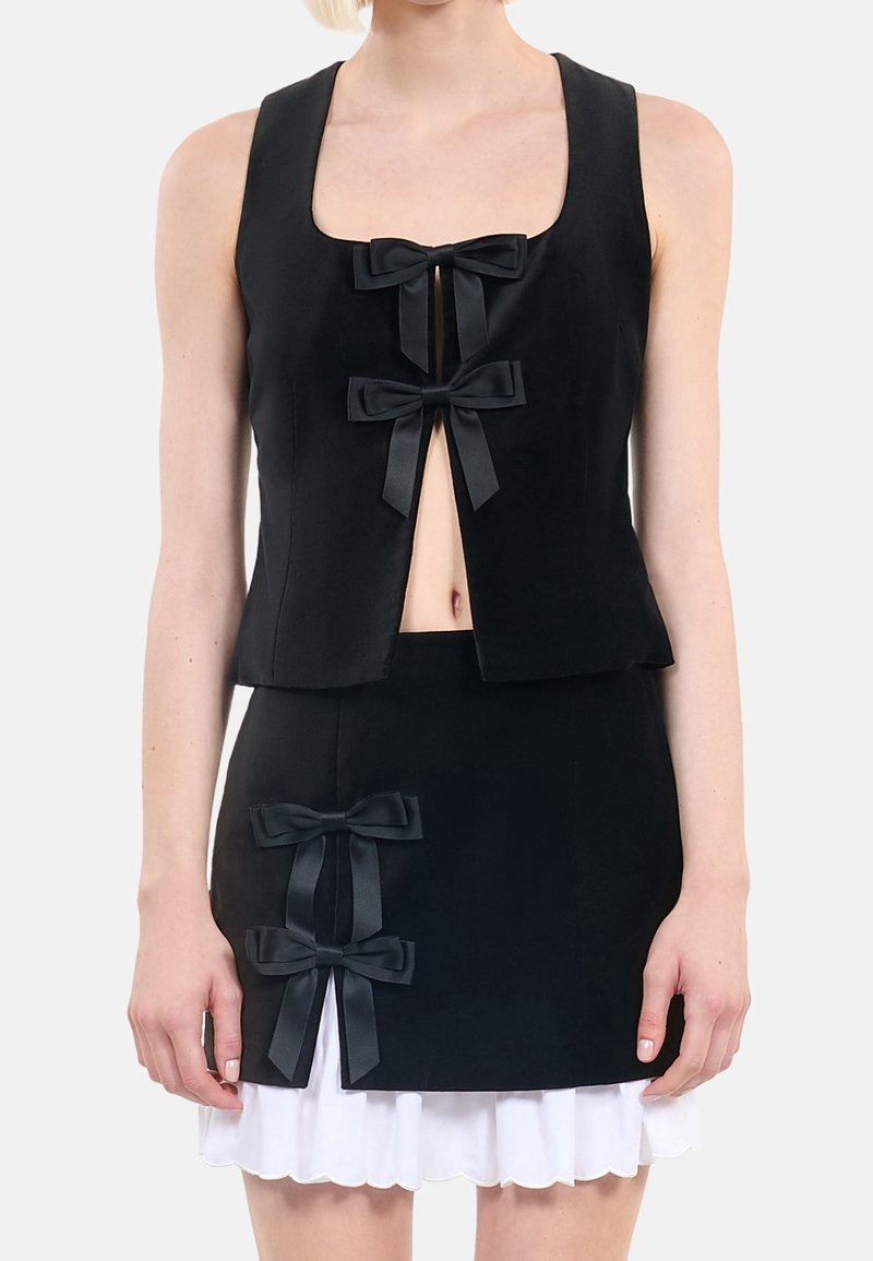 Femme portant un ensemble top sans manches noir et jupe, tous deux ornés de nœuds noirs décoratifs, avec un ourlet blanc à volants visible sous la jupe.