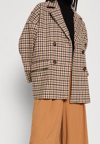Cappotto plaid in beige, marrone e nero, con design doppiopetto, colletto a rever e due tasche anteriori. Tessuto morbido.