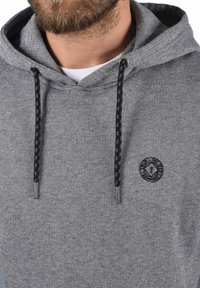 Siva prugasta hoodie s kapuljačom i vezicom, koja ima crne pletene špice i logotip na prsima. Tkanina izgleda teksturirano.