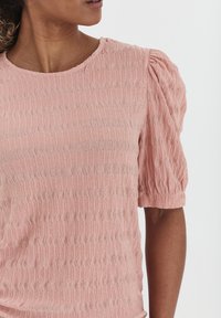 Maglietta rosa chiaro con un motivo testurizzato e ondulato. Presenta un collo a giro e maniche corte a sbuffo. Tessuto morbido con una vestibilità leggermente più rilassata.