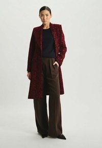 Manteau en velours bourgogne avec une texture florale, design à un bouton, et larges revers ; associé à un pantalon large marron foncé.