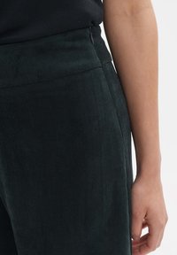 Dark green corduroy pants with a ribbed texture, featuring a high waistband and a side zipper. Close-up of the waist and hand placement.