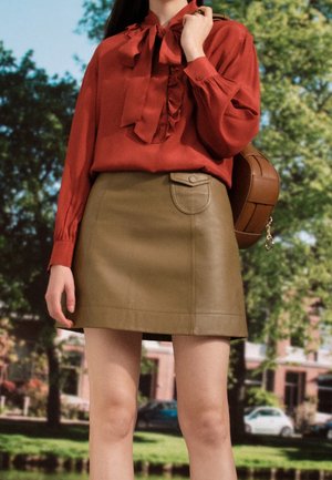 Kobieta ubrana w rdzawobrązową bluzkę z kokardą i brązową skórzaną mini spódniczkę, trzyma brązową torebkę, stoi na zewnątrz na tle drzew.