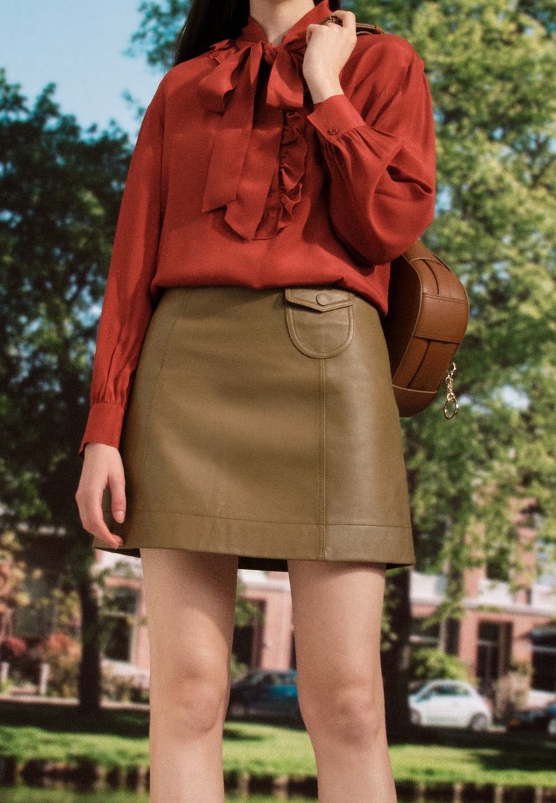 Femme portant un chemisier couleur rouille avec un nœud et une mini-jupe en cuir marron, tenant un sac marron, debout en plein air avec des arbres en arrière-plan.