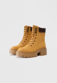 Tan ruskinds ankle boots med en tyk, tekstureret gummisål. Har beige snørebånd, metaløjer og en mørkebrun accent ved kraven.