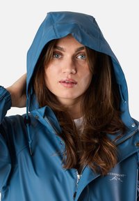 Wodoodporna niebieska kurtka z kapturem, z zamkiem błyskawicznym z przodu oraz sznurkiem do regulacji. Gładka tekstura i logo nadrukowane na klatce piersiowej.