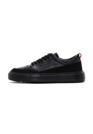 Black sneaker featuring a smooth leather upper with a textured panel, thick rubber sole, and a red accent at the heel. Lace-up design.