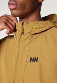 Hnedá zip-up mikina s kapucňou a textúrovaným povrchom. Obsahuje výrazné logo na hrudi a tesný strih okolo krku.