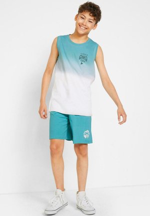 Ragazzo sorridente che indossa una maglietta senza maniche blu e bianca con stampa di un controller di gioco, pantaloni corti teal e sneaker alte bianche.