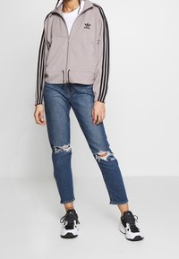 Grå zip-up jacka med svarta ränder, ihop med rivna blå jeans och svarta sneakers. Minimalistisk Adidas-logotyp på jackan.