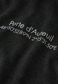 Tessuto grigio scuro con testo bianco stampato che recita "Porte d'Auteuil" e coordinate GPS, con una superficie liscia e leggermente strutturata.