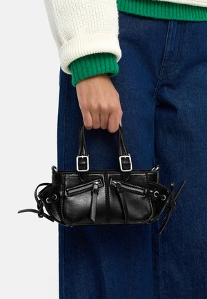 Ręka trzymająca małą czarną skórzaną torebkę z przednimi kieszeniami na suwak, podwójnymi uchwytami oraz bocznymi ozdobami z koronek, na tle niebieskich dżinsów i biało-zielonego swetra.