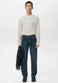 Gestreiftes, langärmliges weißes Hemd, dunkelblaue gerade Jeans, hält einen schwarzen Schal und trägt schwarze Schuhe, steht vor einem hellen Hintergrund.