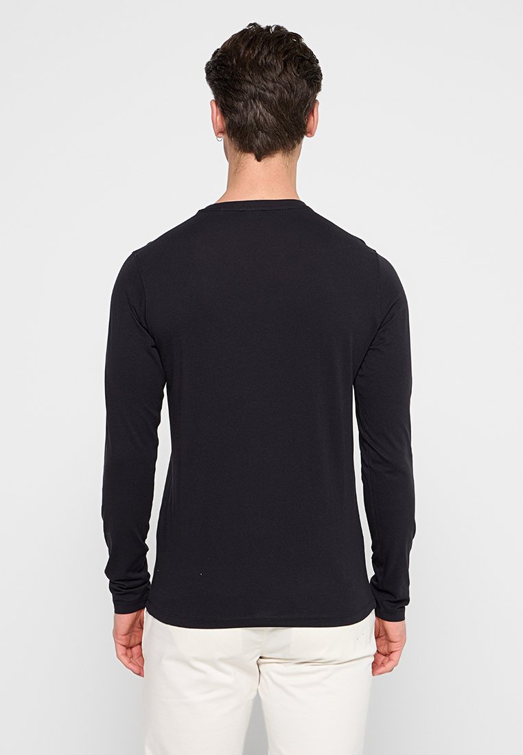 Homme aux cheveux courts et foncés portant une chemise noire à manches longues et un pantalon blanc, se tenant de dos contre un fond clair uni.