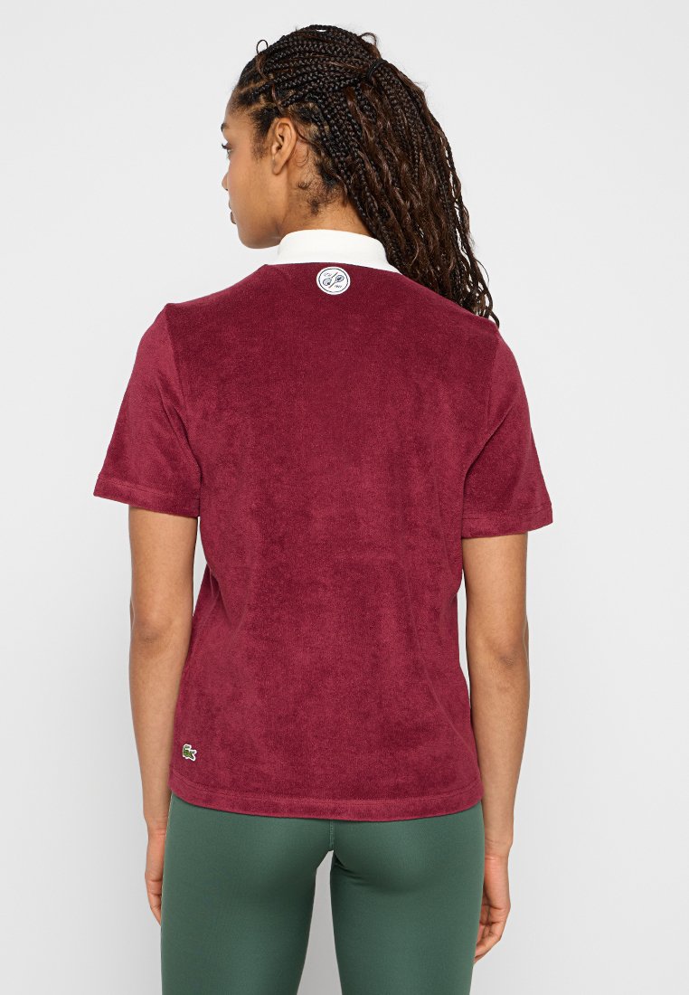 Polo à manches courtes bordeaux avec col, fabriqué en tissu doux texturé, présentant un détail de logo au dos et une petite étiquette de marque sur le côté.