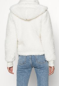 Bílá fleecová bunda s načechranou texturou, kapucí a žebrovanými manžetami. V kombinaci s denimovými džíny se světlejším praním, pohled zezadu.