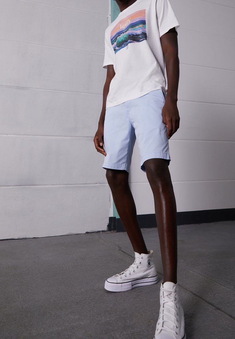 T-shirt graphique blanc avec motif de montagne, associé à un short bleu clair. Porté avec des baskets montantes blanches. Texture lisse, coupe décontractée.