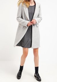Szary wełniany płaszcz z klapowanym kołnierzem, zapięciem na guziki i bocznymi kieszeniami, noszony na ciemnoszarej sukience, zestawiony z czarnymi butami.