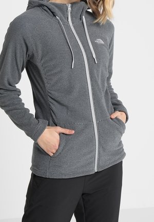 Mujer con chaqueta con capucha gris con cremallera y bolsillos, con las manos en los bolsillos, combinada con pantalones negros sobre un fondo liso.