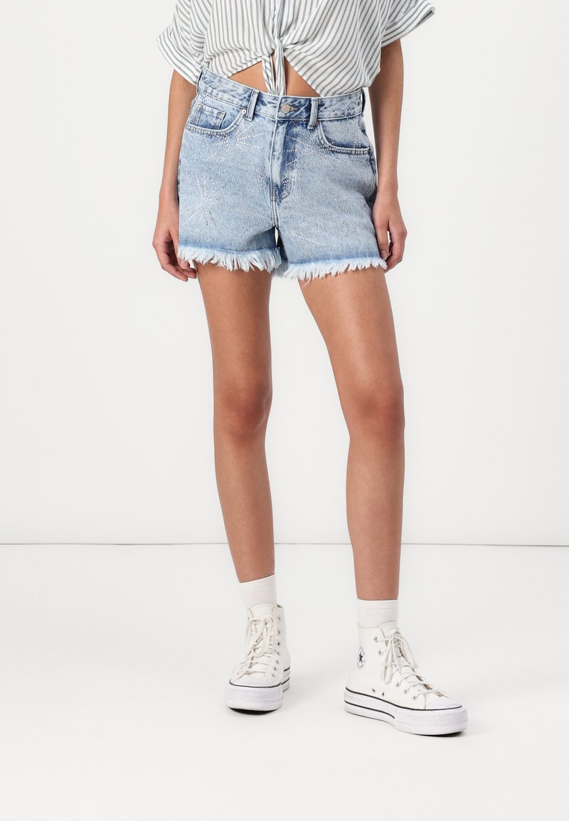 Ljust blå denimshorts med fransad fåll, frampocket och stjärnformade dekorationer, matchat med vita högskurna sneakers och strumpor.
