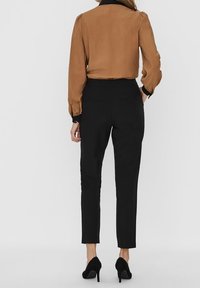 Blusa marrone con maniche a sbuffo, pantaloni neri su misura e scarpe con tacco nere, viste da dietro su uno sfondo neutro.
