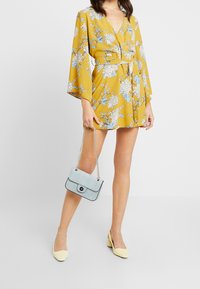 Gelbes Blumenmuster-Kleid mit Glockenärmeln, tailliert und V-Ausschnitt. Trägt eine hellblaue, strukturierte Handtasche mit Kettenriemen.