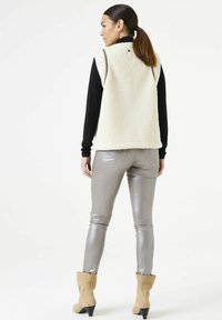 Crème faux fur gilet met hoge kraag, gecombineerd met zilveren leggings en beige enkellaarsjes. Soepele textuur en ontspannen pasvorm. Achteraanzicht.