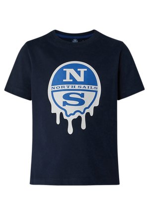 Kind trägt ein dunkelblaues North Sails T-Shirt, schwarze Cargohosen und schwarze Sneakers mit weißen Sohlen, steht mit Händen in den Taschen.
