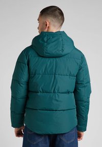 Turkusowa kurtka puchowa z poziomą pikowaną fakturą i kapturem. Posiada elastyczne mankiety i prosty dół. Nosi na sobie niebieskie dżinsy.