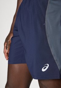 Tamsiai mėlyni sportiniai šortai su šoniniu pilku intarpu, pagaminti iš lengvo audinio. Dėlionėje yra balta logotipas apatinėje priekyje.