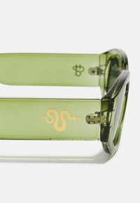 Groene zonnebril met dikke, doorzichtige monturen. Voorzien van een gegraveerd gouden slangembleem op de arm en een metalen scharnierdetail.