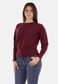 Maglione borgogna con scollatura rotonda, texture a coste e maniche lunghe. Indossato con jeans blu, che evidenziano un design aderente.