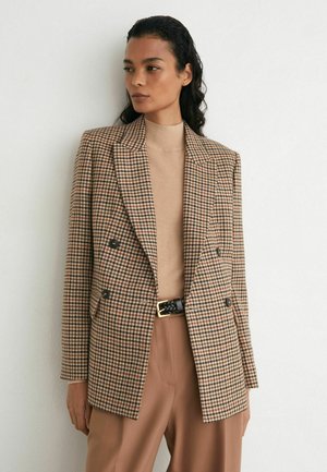 Vrouw die een beige coltrui draagt en een bruin geruit dubbelgeknoopt colbert met brede revers, gecombineerd met hooggetailleerde bruine broek en een zwarte riem.