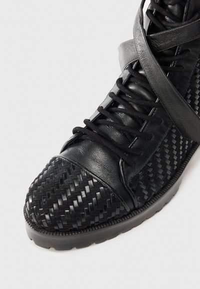 Svart lädersko med flätad struktur på tån, snörning fram och ett brett band över mellanfoten för extra stöd.
