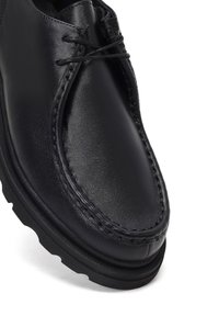 Zapato de cuero negro con un diseño cosido, punta redonda y suela de goma. Los cordones están atados en la parte superior, mostrando una textura suave.