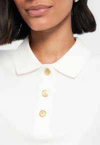 Κοντινό πλάνο ενός ατόμου που φοράει λευκό πουκάμισο με γιακά με τρία χρυσά κουμπιά και έχει κοντά σκούρα μαλλιά ορατά.