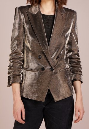 Mujer con blazer metálico bronce de doble botonadura con mangas fruncidas sobre una camiseta negra y pantalones oscuros, torso visible.