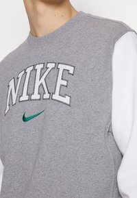 Grå sweatshirt med vita ärmar, med fet "NIKE"-text och en grön swoosh-logotyp. Tillverkad av mjukt, texturerat tyg.