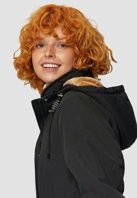 Schwarze Jacke mit strukturiertem Erscheinungsbild, gefütterter Kapuze, verstellbaren Zugbändern und einem Reißverschluss vorne, getragen von einer lächelnden Person mit lockigem orangefarbenem Haar.