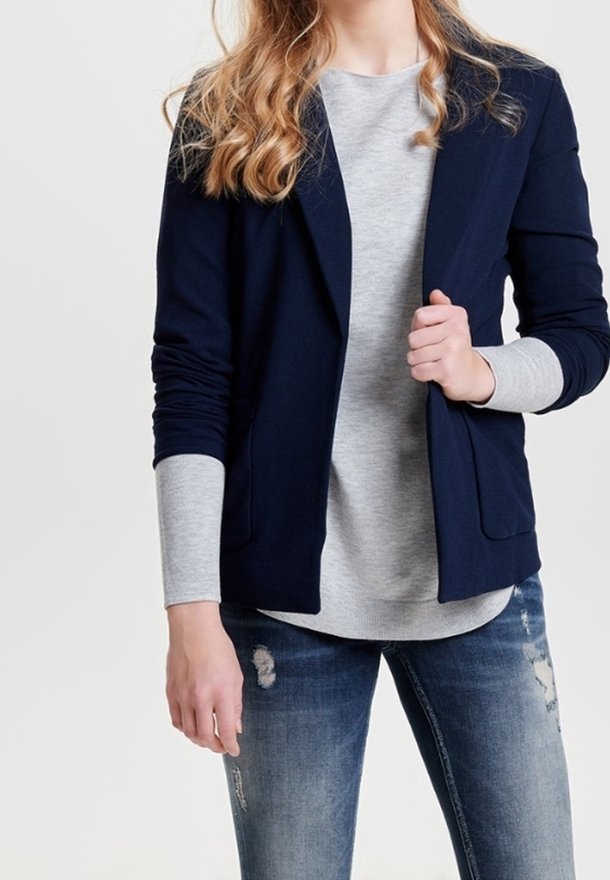 Personne portant un blazer bleu marine sur une chemise grise à manches longues et un jean bleu déchiré, posant avec une main tenant le blazer.