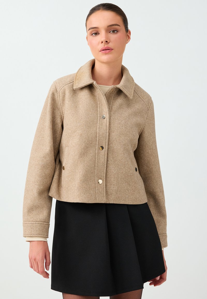 Woman wearing a beige buttoned wool jacket with a collar and a black pleated skirt against a plain light background.