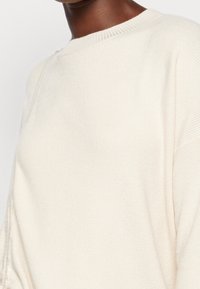 Pull en maille côtelée douce de couleur crème clair avec un col rond large et des épaules tombantes. Texture lisse avec un minimum de détails.