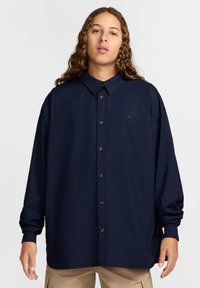 Camisa azul marinho de botão com colarinho, apresentando um pequeno logótipo bordado, mangas longas e botões castanhos, desenhada com um corte descontraído.