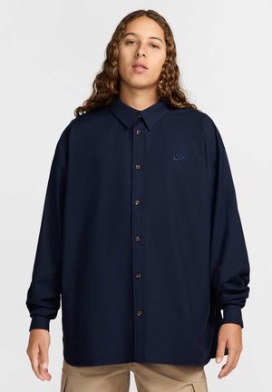 Chemise bleu marine à boutons avec col, arborant un petit logo brodé, manches longues et boutons marron, conçue avec une coupe décontractée.
