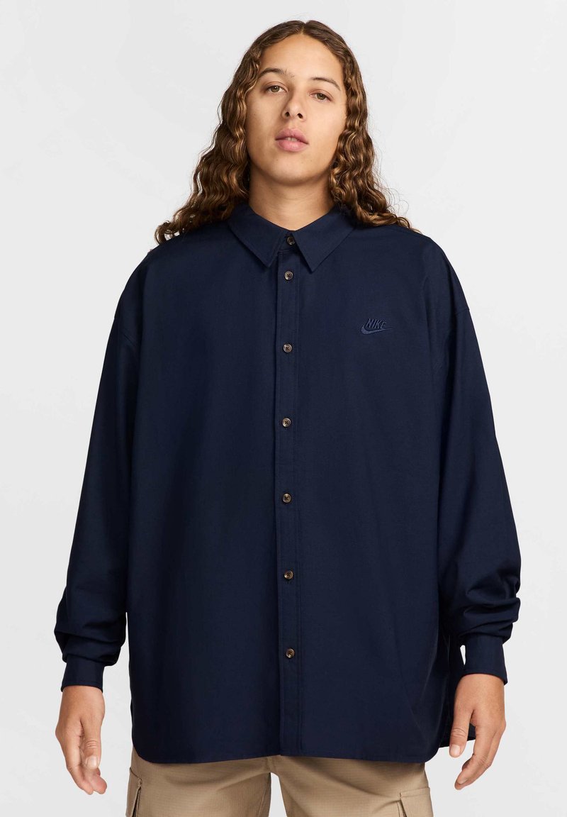 Camisa azul marinho de botão com colarinho, apresentando um pequeno logótipo bordado, mangas longas e botões castanhos, desenhada com um corte descontraído.