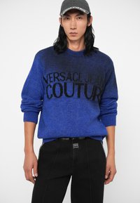 Kék kötött pulóver, amelyen fekete "VERSACE JEAN COUTURE" felirat található, bordázott mandzsettákkal és aljjal, fekete texturált farmernadrággal párosítva.