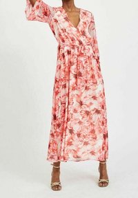 Kvinne iført en lang, luftig, rosa og rød blomstrete omslagskjole med 3/4 ermer og beige høye sandaler, stående mot en ensartet bakgrunn.