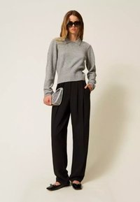 Maglione grigio lavorato a maglia con colletto, pantaloni neri plissettati e ballerine nere. In mano tiene una piccola pochette con motivo. Sfondo semplice.